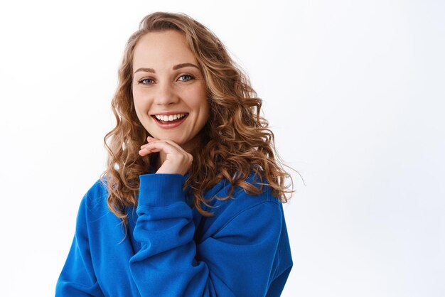 Jolie femme blonde avec une coiffure bouclée touchant le menton et souriant ravie de regarder la caméra avec intérêt et joie debout sur fond blanc