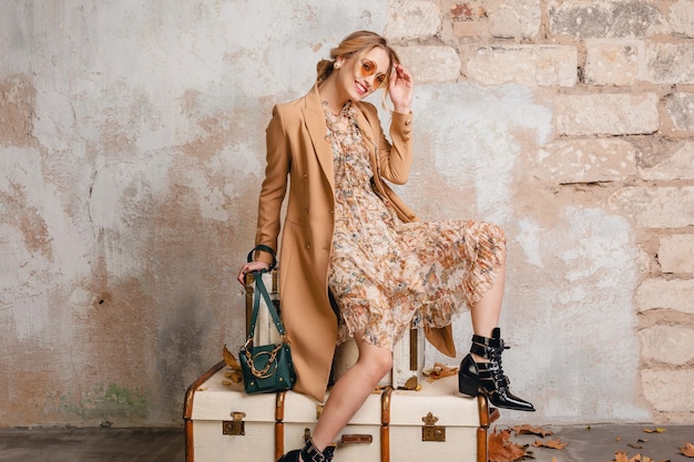 Jolie femme blonde élégante en manteau beige assis sur des valises contre le mur dans la rue
