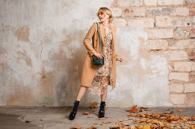 Jolie femme blonde élégante en manteau beige marchant dans la rue contre le mur vintage