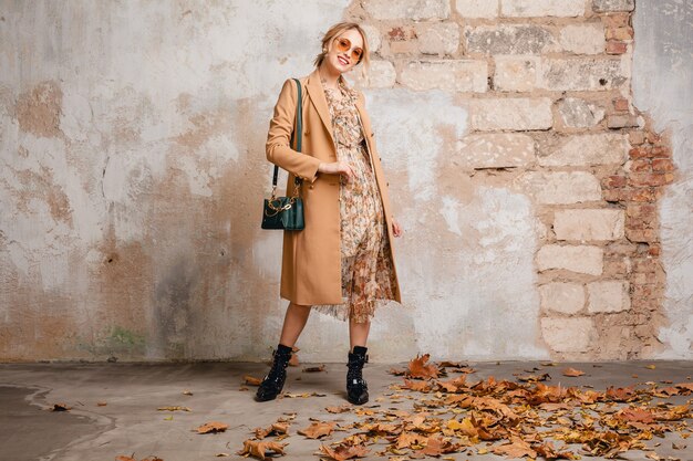 Jolie femme blonde élégante en manteau beige marchant dans la rue contre le mur vintage