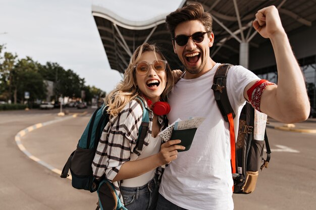 Jolie femme blonde à lunettes de soleil et homme en tee blanc sourit et prend selfie près de l'aéroport