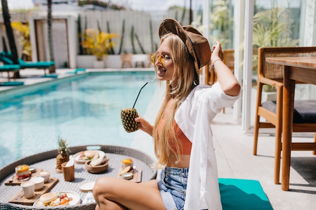 Jolie femme blonde en lunettes de soleil jaunes, passer du temps près de la piscine avec un délicieux cocktail d'ananas. Touriste rêveuse mangeant des fruits pendant la séance photo à la station.