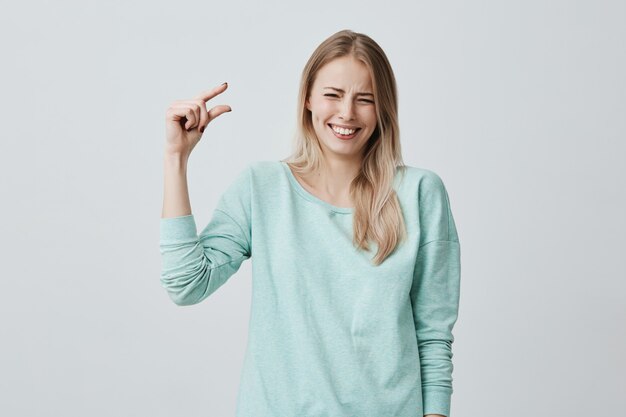 Jolie femme blonde mignonne dans des vêtements décontractés montrant quelque chose de petite taille avec les mains tout en gesticulant, souriant largement avec des dents. Blonde européenne femelle démontrant la taille de la petite boîte