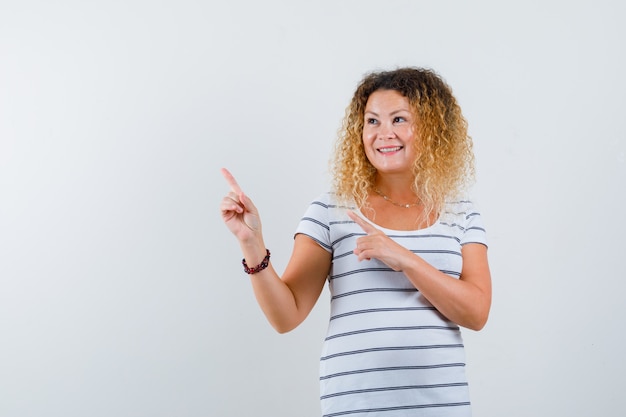 Jolie femme blonde pointant vers le coin supérieur gauche en t-shirt rayé et l'air joyeux. vue de face.