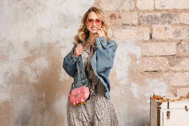 Jolie femme blonde souriante élégante en jeans et veste surdimensionnée marchant contre le mur dans la rue