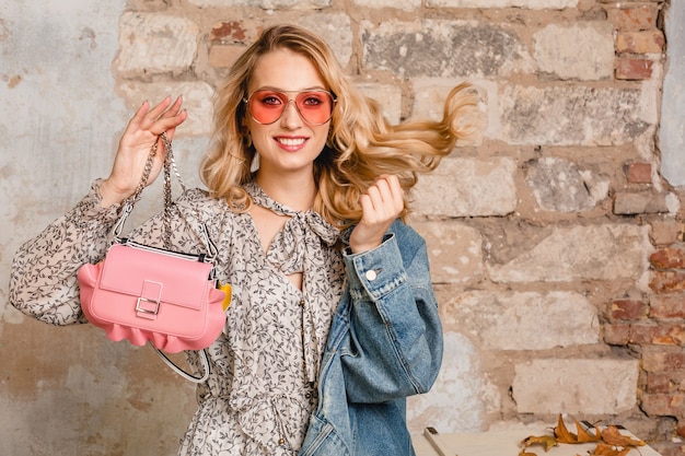 Jolie femme blonde souriante élégante en veste de jeans marchant contre le mur dans la rue