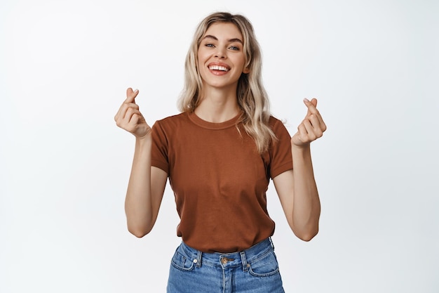 Jolie femme blonde souriante montrant le geste des coeurs des doigts debout dans des vêtements décontractés sur fond blanc