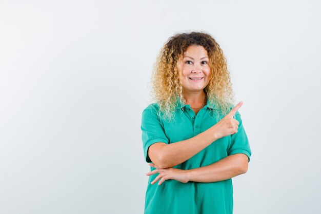 Jolie femme blonde en t-shirt de polo vert pointant vers le coin supérieur droit et à la vue de face, joyeuse.