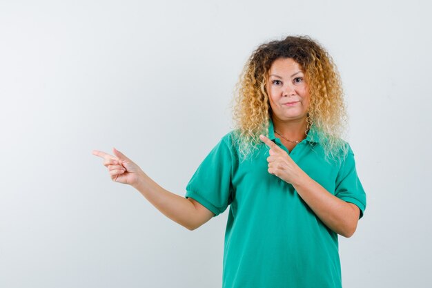 Jolie femme blonde en t-shirt de polo vert pointant vers le coin supérieur gauche et regardant hésitante, vue de face.