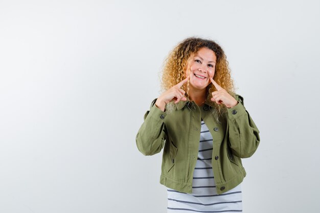 Jolie femme blonde en veste verte pointant sur son sourire et à la joyeuse vue de face.