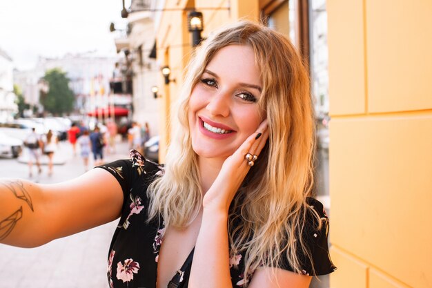 Jolie femme blonde vêtue d'une robe à fleurs, posant près du mur jaune