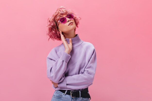 Jolie femme bouclée en pull en cachemire violet et lunettes de soleil fuchsia pose sur un mur isolé