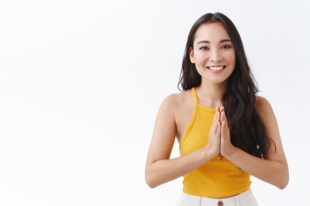 Jolie femme brune d'Asie de l'Est en haut jaune tendance, serrer les mains ensemble en namaste, mendier ou prier, souriant sans soucis, regarde avec gratitude, remercie de l'aide, fond blanc