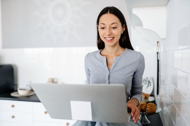 Jolie femme brune assise sur la table à la cuisine et à l'aide d'un ordinateur portable