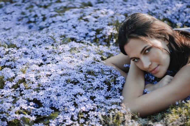 Photo gratuite jolie femme brune au look sexy est allongée sur la cour avec des fleurs bleues