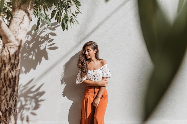 Jolie femme brune aux cheveux longs en jupes-culottes élégantes et haut blanc posant contre un mur blanc et un olivier