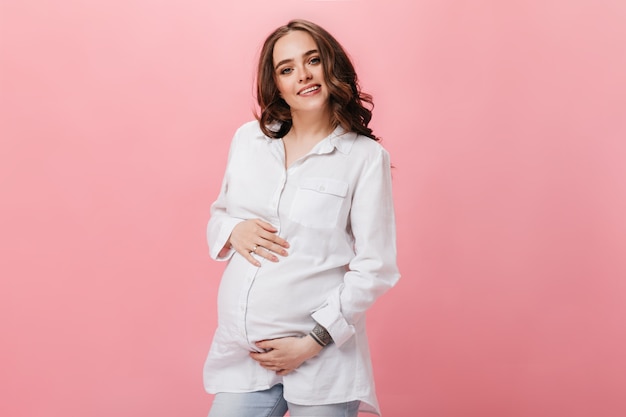 Jolie femme brune en chemise blanche et jeans sourit et touche le ventre. Fille enceinte en jeans pose sur fond rose.