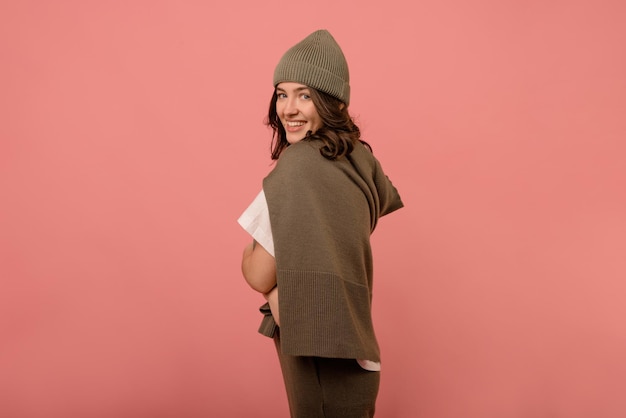 Jolie femme brune européenne de 20 ans en chapeau et veste regarde la caméra par-dessus l'épaule sur fond rose Concept de loisirs de style de vie