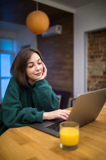 Jolie femme brune heureuse travaille sur son ordinateur portable sur la table de la cuisine, boire du jus d'orange