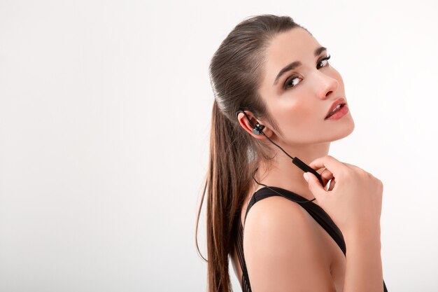 Jolie femme brune en jogging haut noir, écouter de la musique sur les écouteurs posant isolé sur la coiffure de queue de cheval de mur blanc