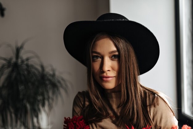 Jolie femme brune à la peau bronzée se penche sur la caméra. Femme au chapeau noir et t-shirt beige pose dans une pièce lumineuse.