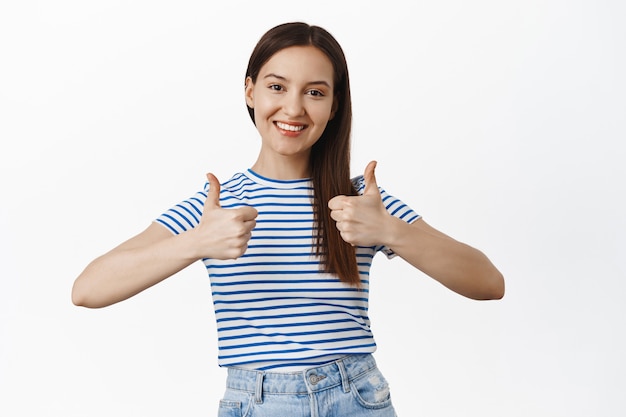 Photo gratuite une jolie femme brune souriante montre son pouce levé, hoche la tête en signe d'approbation, recommande la vente, loue un bon choix, un excellent travail, satisfait de smth, aime et accepte, mur blanc.