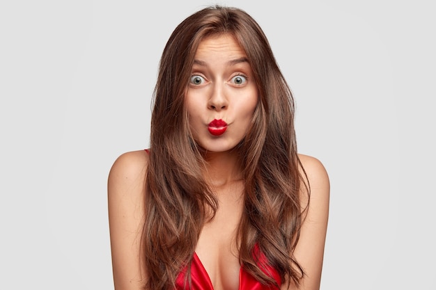 Jolie femme brune surprise garde les lèvres rouges arrondies, a les yeux sortis, une peau pure, voit quelque chose d'incroyable devant, pose pour un magazine de mode, isolé sur un mur blanc. Concept de beauté