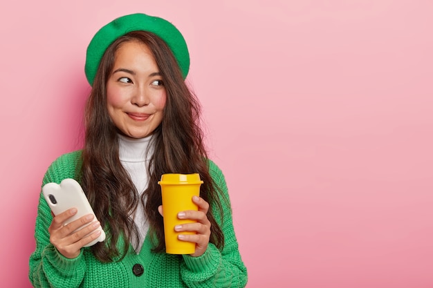 Jolie femme brune en tenue verte, tient le téléphone portable dans une main et une tasse de café dans d'autres regards avec une expression de visage de rêve