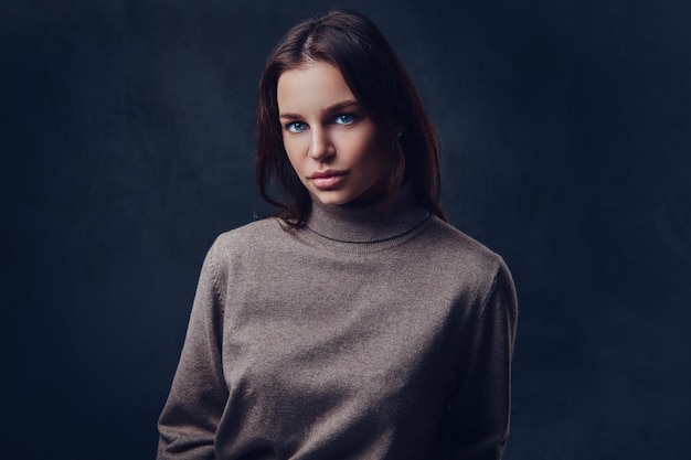 Une jolie femme brune vêtue d'une veste marron à long cou sur fond gris.