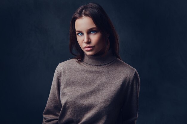 Une jolie femme brune vêtue d'une veste marron à long cou sur fond gris.