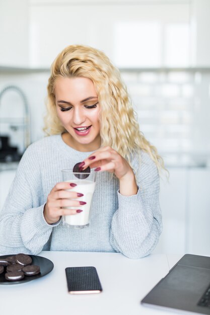 Jolie femme buvant du lait dans un verre, mangeant des cookies à l'aide d'un ordinateur portable tout en étant assis à la table de la cuisine