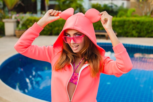 Jolie femme à capuche rose coloré portant des lunettes de soleil sur les vacances d'été souriant expression du visage émotionnel s'amuser, style de mode sport