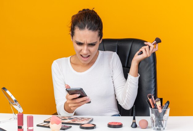 Jolie femme caucasienne agacée assise à table avec des outils de maquillage tenant un pinceau de maquillage et regardant le téléphone