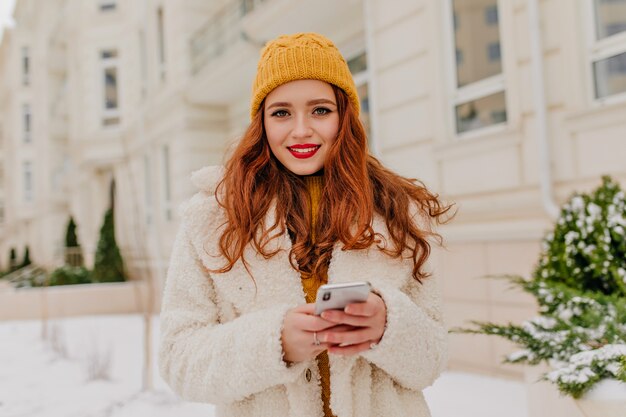 Jolie femme caucasienne en bonnet tricoté tenant le téléphone. Photo extérieure d'une fille au gingembre inspirée portant un manteau blanc.