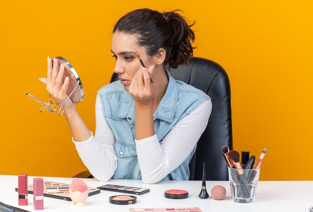 Jolie femme caucasienne confiante assise à table avec des outils de maquillage regardant un miroir appliquant un fard à paupières