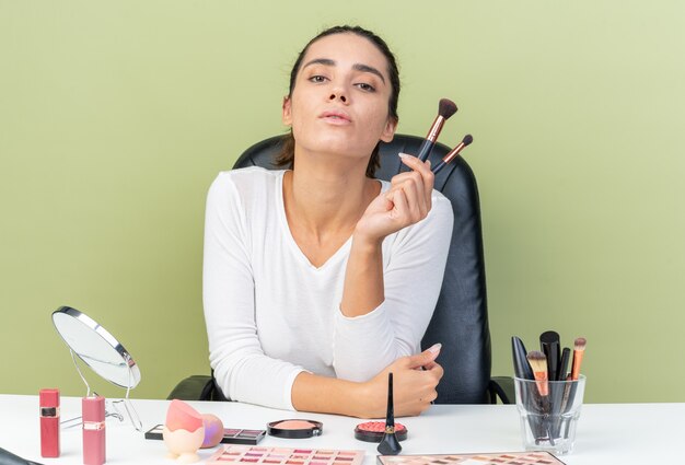 Jolie femme caucasienne confiante assise à table avec des outils de maquillage tenant des pinceaux de maquillage