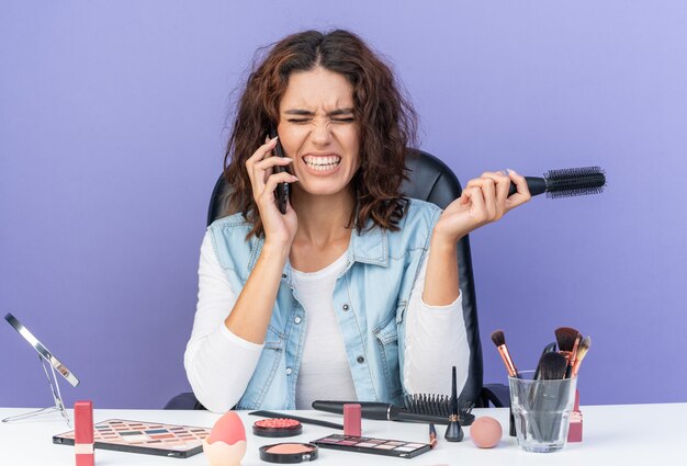 Jolie femme caucasienne mécontente assise à table avec des outils de maquillage parlant au téléphone et tenant un peigne isolé sur un mur violet avec espace de copie