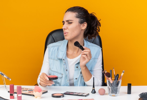 Jolie femme caucasienne mécontente assise à table avec des outils de maquillage tenant un pinceau de maquillage et rougir en regardant le côté isolé sur un mur orange avec espace de copie