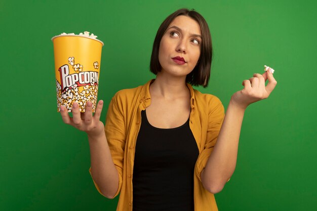 Jolie femme caucasienne réfléchie tient un seau de pop-corn et lève les yeux sur le vert