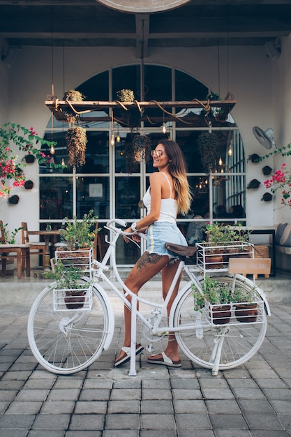 Jolie femme caucasienne tatouée en short en jean et haut blanc se dresse à vélo sur fond de café de rue.