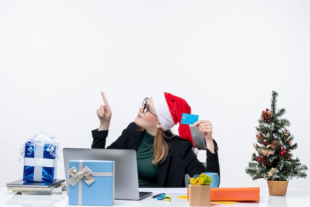 Jolie femme avec chapeau de père Noël et portant des lunettes assis à une table cadeau de Noël et tenant une carte bancaire à la recherche au-dessus dans le bureau