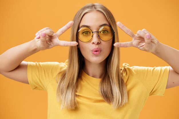 Jolie femme charismatique insouciante dans des lunettes de soleil rondes à la mode et un t-shirt pliant les lèvres dans un baiser montrant des signes de paix ou de disco près des yeux dansant en s'amusant à la fête sur fond orange. Mode de vie.