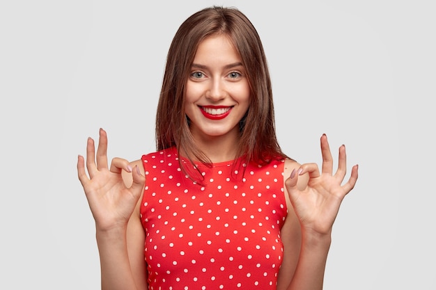 Jolie femme avec un charmant sourire amical, fait un geste correct, vêtue d'une robe à pois à la mode, montre son approbation, pose contre un mur blanc. Jeune femme avec des modèles de lèvres rouges à l'intérieur.