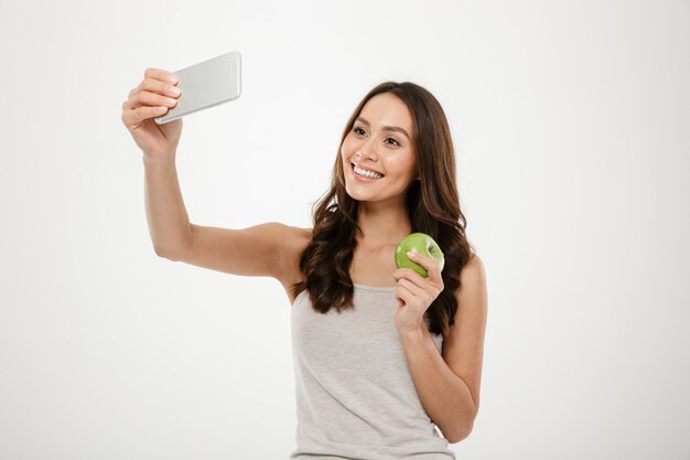 Jolie femme charmante faisant selfie sur téléphone portable argent et tenant la pomme verte juteuse, isolé sur mur blanc