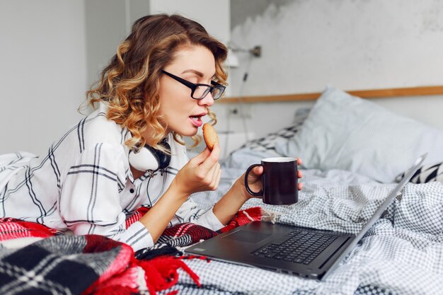 Jolie femme avec une coiffure ondulée, manger des cookies avec du café, regarder la vidéo sur son ordinateur portable.