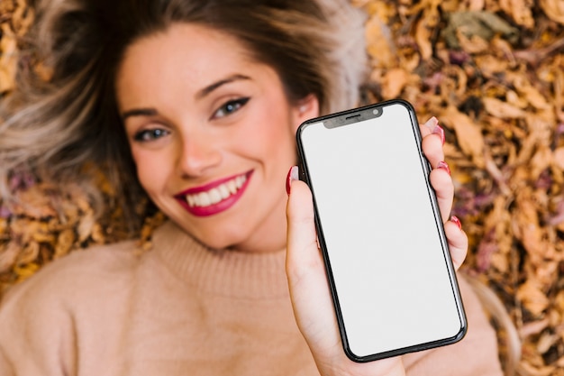 Jolie femme couchée sur des feuilles sèches montrant un téléphone portable à écran blanc