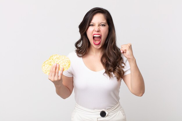 Jolie femme criant agressivement avec une expression fâchée et tenant des gâteaux de riz de régime