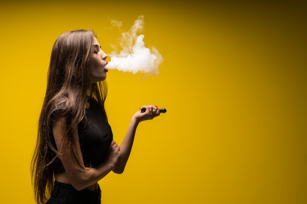 Jolie femme debout et vaping sur le mur jaune.