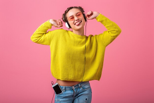 Jolie femme drôle heureuse, écouter de la musique dans des écouteurs habillés en tenue de style coloré hipster isolé sur un mur rose, portant un pull jaune et des lunettes de soleil, s'amuser