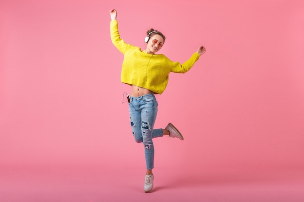 Jolie femme drôle heureuse en pull jaune sautant à écouter de la musique dans des écouteurs habillés en tenue de style coloré hipster isolé sur mur rose, s'amuser
